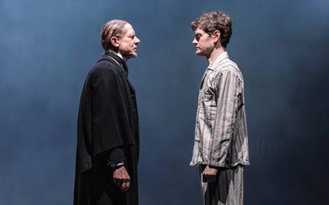 Jerry Killick wears a black overcoat and stands in front of Kyle Soller, who wears blue stripey pyjamas. They face each other.
