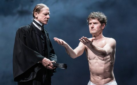 Jerry Killick wears a black overcoat and holds a belt. He has a menacing expression and stands to the side of Kyle Soller, who is topless and holds his arms out in front of him.