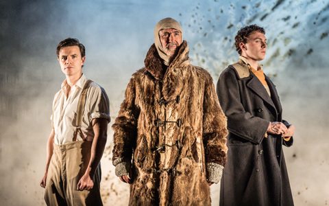 Declan Conlon wears a large fur coat and woollen head covering. He looks ahead, with Oliver Johnstone and Stuart Thompson standing on each side of him.