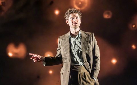 Kyle Soller wears a brown striped suit jacket and gestures to the side with one hand.