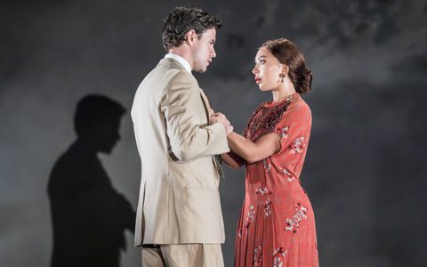 Oliver Johnstone wears a light beige suit and stands holding Yanexi Enriquez’s hands. She wears a red dress. They look at one another.