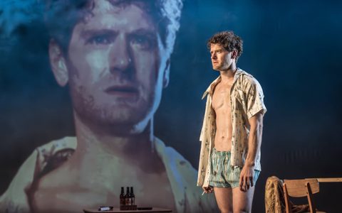 Kyle Soller wears blue shorts and a loose open shirt. He stands in front of a table, holding a cigarette. A close-up of his face, with a serious expression, is projected behind him.