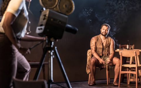 Olivier Huband wears green shorts and a brown fringed waistcoat. He sits on a wooden outdoor table and looks towards Adelle Leonce. She is blurred in the foreground, manoeuvring a camera on a tripod.