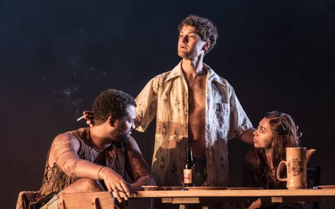 Kyle Soller wears a loose open shirt and looks off to the side. Yanexi Enriquez and Olivier Huband sit a wooden outdoor table either side of Kyle, looking towards him. He stands with his hands on their shoulders.