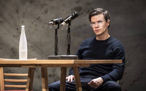 Oliver Johnstone wears a black crewneck and sits at a table in a wide, confident manner. There are microphones and a bottle of water on the table in front of him.