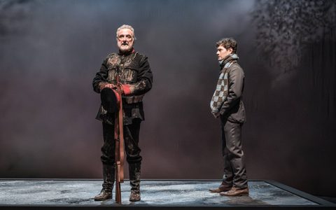 Declan Conlon wears a black and red army uniform. He looks ahead, holding a rife. Kyle Soller wears a blue and black scarf and stands to the side, looking at him.