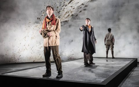 Kyle Soller wears a dark beige jacket and a yellow and red scarf. He looks up, and Stuart Thompson wears a dark coat and stands mid stage, gesturing towards Kyle. Oliver Johnstone stands at the back, facing away from the camera.