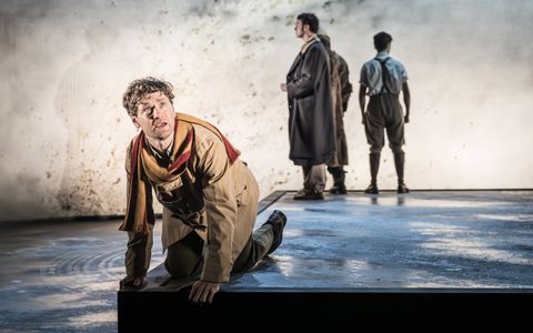 Kyle Soller wears a dark beige jacket and a yellow and red scarf. He kneels at the edge of the stage and looks up with a frightened expression. Stuart Thompson, Oliver Johnstone and Declan Conlon stand in a corner in the background.
