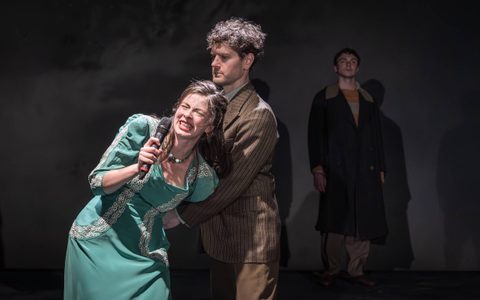 Agnes O’Casey wears a teal dress and holds a microphone. She cries as Kyle Soller restrains her arms from behind. He wears a brown striped suit jacket.