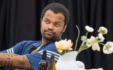 Olivier Huband wears a blue t-shirt and sits at a table. He has a neutral expression and on the table are glass bottles and flowers in a vase.
