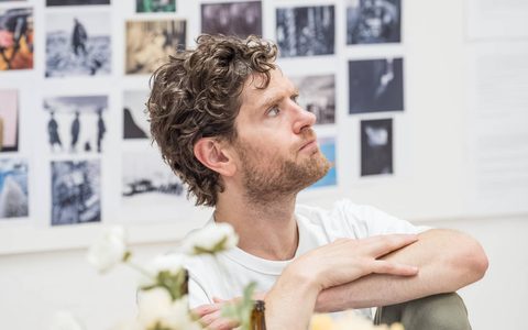 Kyle Soller wears a white t-shirt and sits at a table, resting his arms on his knee. He looks off to the side with a thoughtful expression. On the table are glass bottles and flowers in a vase.