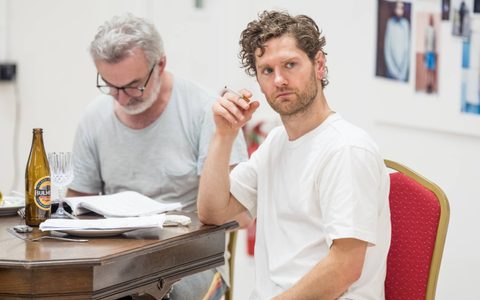 Kyle Soller wears a white t-shirt and sits a wooden table. He looks off to the side and holds a cigarette in his hand. Declan Conlon sits at the table in the background, looking down at a script.
