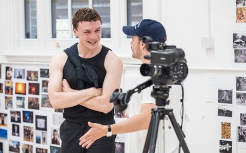 Stuart Thompson wears a black tank top and smiles with his arms crossed. He is in conversation with Oliver Johnstone, who wears a white t-shirt and a blue cap. There is a camera on a tripod in the foreground, partially obscuring Oliver.