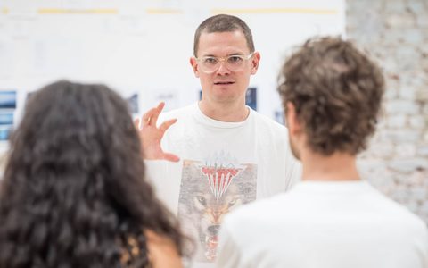 Sam Pritchard wears a white t-shirt and gestures with one hand. Adelle Leonce and Kyle Soller are in the foreground, facing towards Sam.