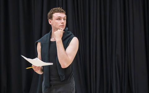 Stuart Thompson wears a black tank top with a black sweater over his shoulders. He looks off to the side with one hand on his chin, holding a script in the other hand.
