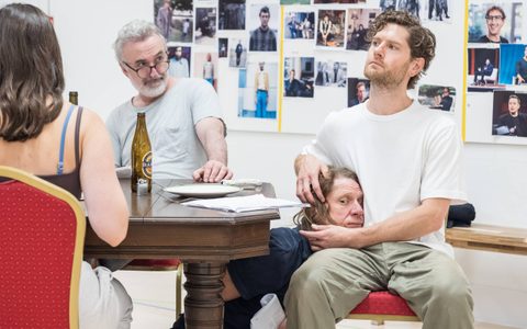 Jerry Killick sits underneath a wooden table, resting his head against Kyle Soller, who wears a white t-shirt and sits at the table. Declan Conlon looks towards them in the background.