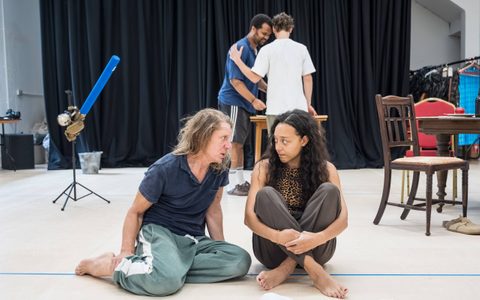 Adelle Leonce sits on the floor with her legs crossed, talking to Jerry Killick, who wears a blue t-shirt. She has a worried expression. Olivier Huband and Kyle Soller stand in the background with their backs to the camera.