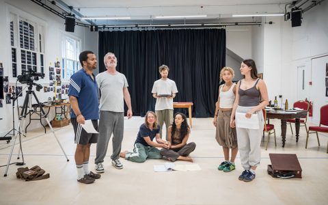 The Romans company gathered in a rehearsal room. They stand in a semi-circle holding scripts, with Adelle Leonce and Jerry Killick sitting on the floor in the middle.