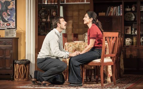 Leila Farzad sits at a dining room table smiling up towards the ceiling. Nicholas Karimi kneels on the ground in front of her and holds her hand.