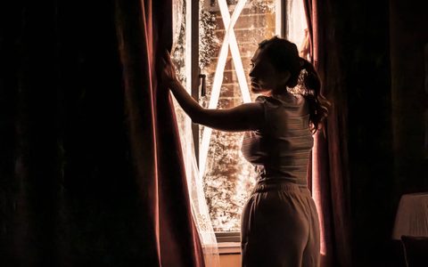 Leila Farzad stands in front of a taped up window, with her hands pulling both of the curtains apart. The room is dark, and the only light pours in through the window.