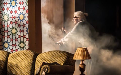 Souad Faress stands with her left hand on the handle of an open door. Through the door pours white smoke.