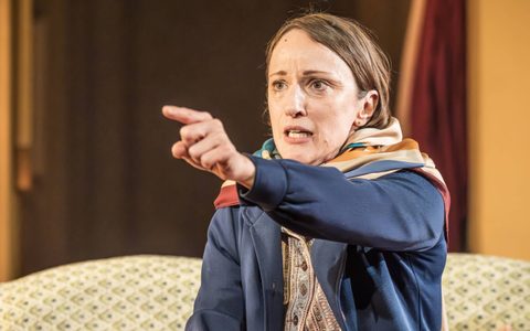 Nadia Albina sits on a sofa, wearing a blue jacket. She points forward with her left hand, she has a pained expression on her face.