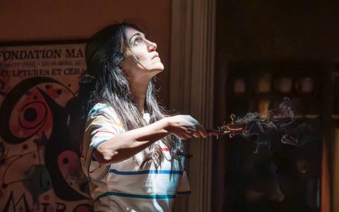 Leila Farzad stands in a dark room, illuminated by a beam of bright light. She is holding efsand seeds, which are burning.