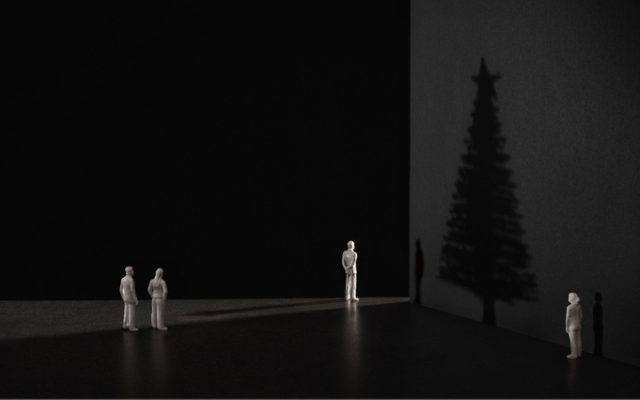 The shadow of a Christmas tree against a dark wall, with four small white figurines looking at it. 