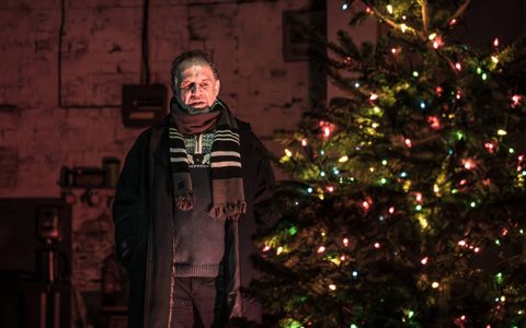 Nigel Lindsay wears a big black coat and a stripy scarf on stage. He looks disdainfully at a Christmas tree.