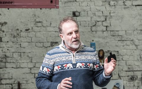 Nigel Lindsay wears a blue Christmas winter-themed sweater. He stands by a dining table on stage, gesturing with his hands.