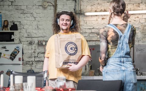 Jamie Ankrah wears a yellow t-shirt and over the ear headphones. He smiles at Callie Cooke, who has her back to the camera. He holds a chicken shop paper bag.