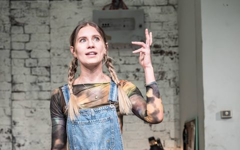 Callie Cooke wears blue dungarees and stands at a dining table laid with candles. She speaks, gesturing with one hand.