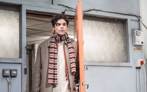 Jacob Fortune-Lloyd stands in an orange and grey doorway with one hand on the handle. He wears a grey coat and stripey scarf.