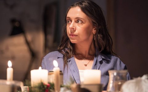 Bel Powley wears a blue striped shirt and sits at a dining table. She looks off to the side.