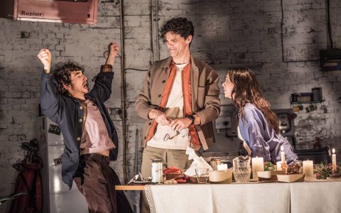 Samuel Blenkin, Jaco Fortune-Lloyd and Bel Powley stand behind a dining table on stage. Samuel gestures wildly as Jacob and Bel face him. The table is laden with food containers and drinks.