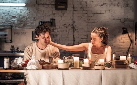 Bel Powley sits next to Jacob Fortune-Lloyd at a dining table laden with food containers. She rests one hand on Jacob’s face, and both look down.