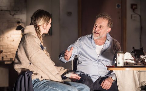 Nigel Lindsay and Callie Cooke sit at a dining table on stage. Nigel gestures with one hand as they talk.