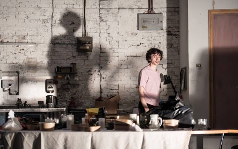 Samuel Blenkin looks towards a dining table on stage, which is laden with food containers. The stage is a dilapidated room.