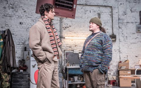 Jacob Fortune-Lloyd and Jamie Ankrah stand talking on stage. The set is a dilapidated room with white brick walls.