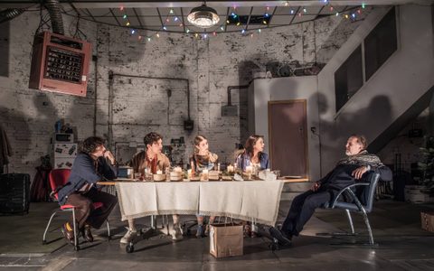 The Christmas Day company sit around a dining table laden with takeaway containers, drinks and candles. The stage is a dilapidated room with white brick walls and a fan heater hanging overhead.
