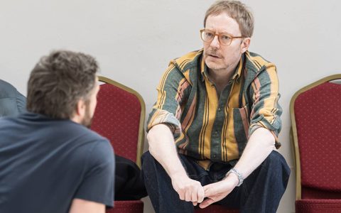 Director Joe Hill-Gibbins wears a green and orange shirt and sits on a red chair.