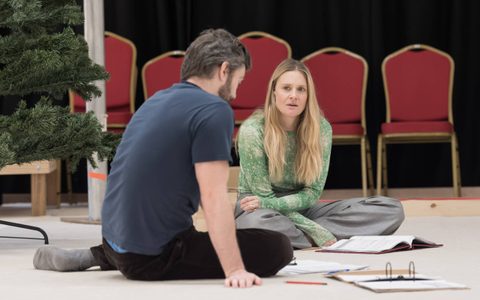 Romola Garai sits crossed-legged on the floor and looks at James Corrigan who sits opposite her with his back to us.