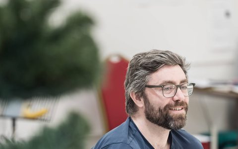 James Corrigan wears glasses and is seen through branches of a Christmas tree as he smiles.