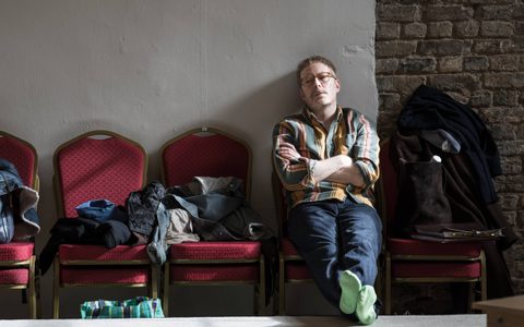 Director Joe Hill- Gibbins sits with his arms crossed on a row of red chairs beside a pile of clothes and bags, leaning against a wall.