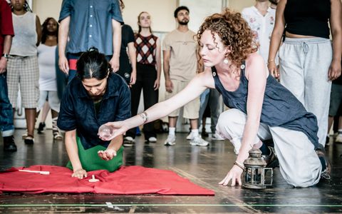 Two women crouch on a red mat on the floor. One holds a spherical glass globe up to the other.