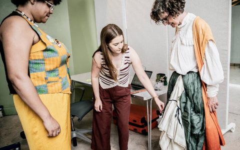 Three people gather around a woman, looking at the dress she is wearing.