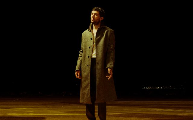 Josh O'Connor stands on a yellow floor, against a black background. His eyes are closed, and his left hand is partially splayed.