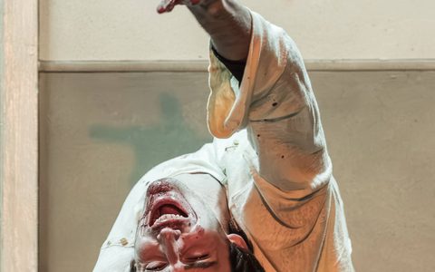 Luke Cinque-White backbends with his right hand over his head. He has blood around his mouth and he screams with his eyes open.