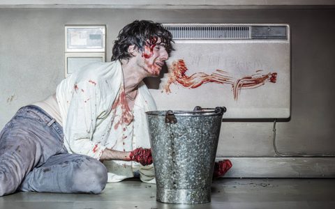 Luke Cinque-White leans against a blood-smeared radiator on the floor. There is a silver bucket in front of him. He hands are wrapped in rope and covered in blood.