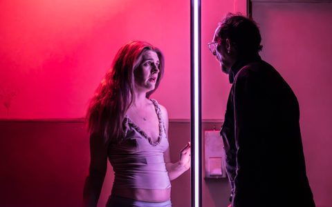 Lizzy Connolly and Leo Bill look at each other through a large window. The lighting is pink.